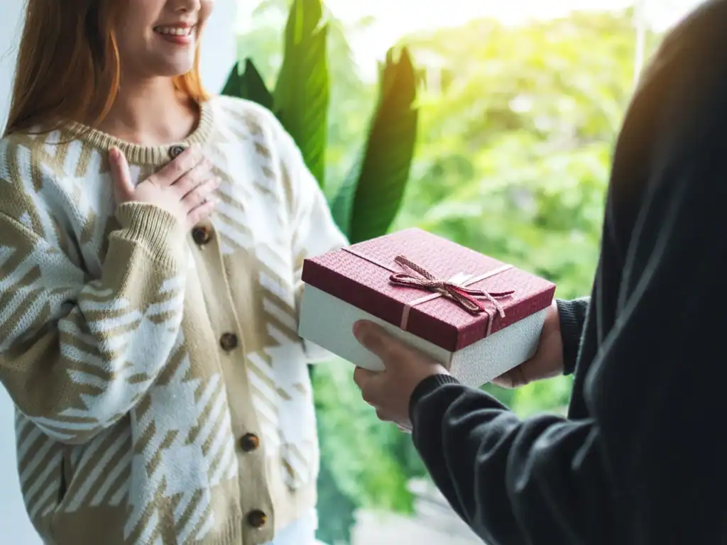 Surprise gift exchange for a loved one, woman happily receiving a wrapped present, indoors with lush greenery outside.