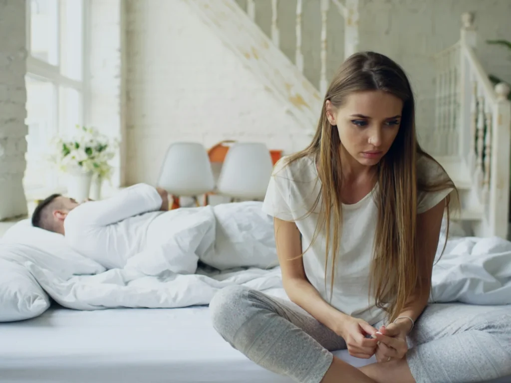 Woman sitting on the edge of a bed while her spouse sleeps, feeling disconnected